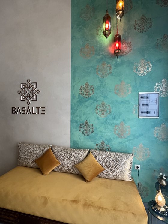 Le mur signature Basalte : l'emblème et le nom sur enduit sable, à côté d'un mur eucalyptus à motifs et de lanternes marocaines