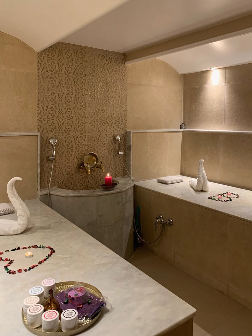 La salle de hammam : murs en tadelakt et marbre, robinetterie en laiton, serviette pliée en cygne et pétales de rose