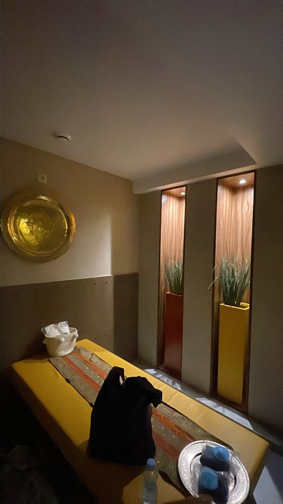 La salle de massage : table drapée de mustard, disque en laiton au mur et niches en bois plantées de graminées